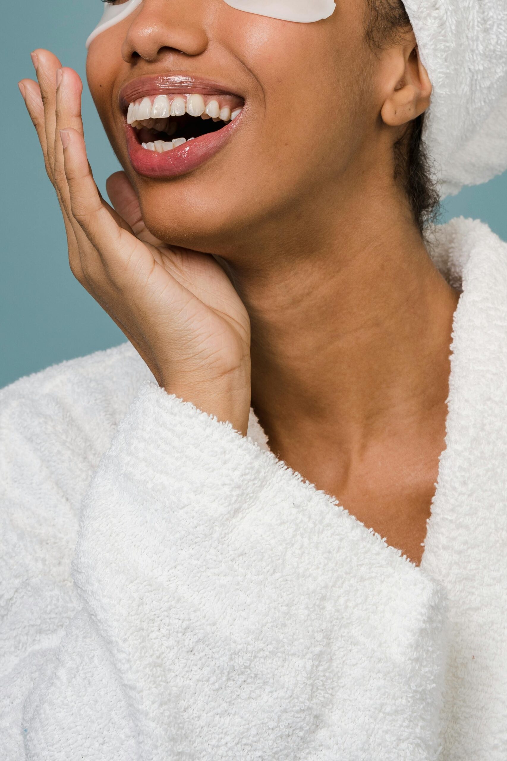 Crop unrecognizable young ethnic female in bathrobe withe eye patches and tower on head laughing and touching perfect face during against blue background